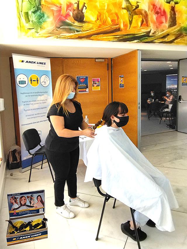 Όρθια η Διευθύντρια των κομμωτηρίων Art & Beaty, Κατερίνα Μπιτσάκη λίγο πριν κόψει από νεαρή εθελόντρια μία ακόμα κοτσίδα, η οποία προορίζεται για περούκες καρκινοπαθών του Συλλόγου "ΕΥ ΖΩ".